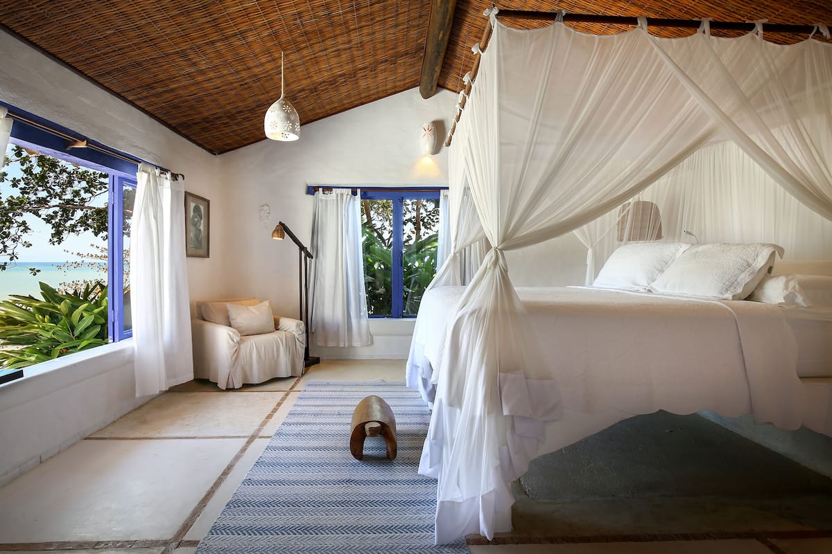 A bedroom with a canopy bed, a view of the sea, and a small wooden stool.