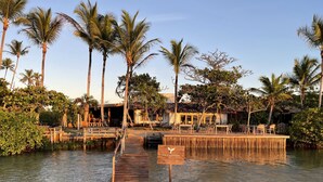 Un muelle de madera que lleva a un edificio con mesas al aire libre, rodeado de palmeras y agua.