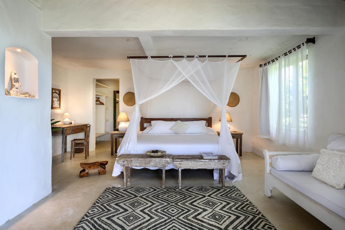 A bedroom with a four-poster bed, a wooden bench, a white sofa, and a patterned rug.