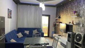 A living room with a blue sofa, a dining table, and a TV. There is a window with curtains, a fan, and a refrigerator. - Luxury Apartment in ALDAU HEIGHTS 10204 (Hurghada)