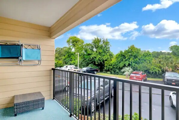 A balcony with a metal railing, a wicker bench, and a view of parked cars and greenery. - 3 min walk to Folly Field Beach Modern & Fun 1bdrm (Hilton Head Island)