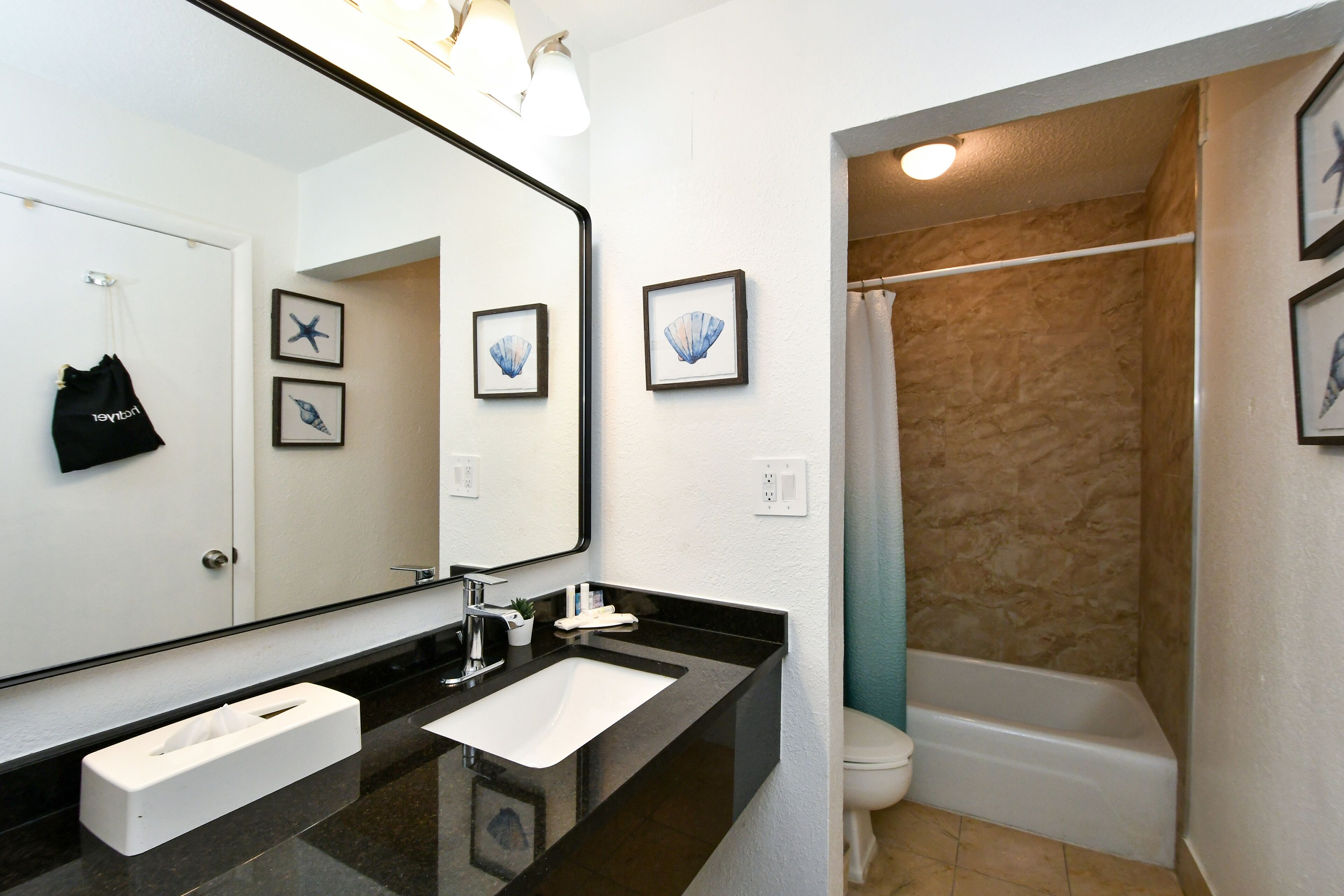 A modern bathroom with a black countertop, white sink, and a large mirror. There are framed pictures on the wall and a black bag hanging on the door.