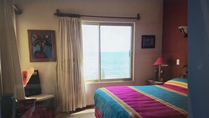 Un dormitorio con cama, ventana con vista al mar, una silla, una lámpara y un cuadro en la pared.