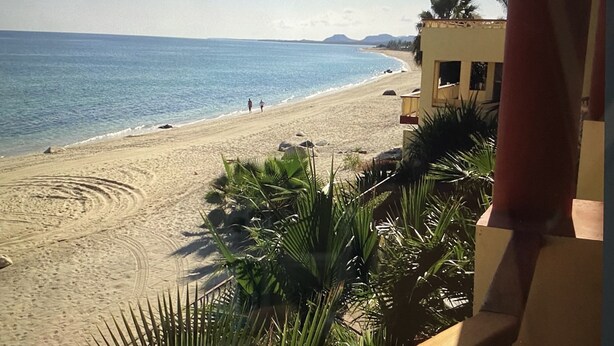 Una playa con arena dorada, mar azul cristalino y algunas personas caminando. Se observa un edificio con balcón que da al mar.