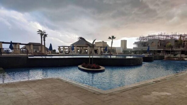 A swimming pool area with a tiled deck, a central water feature, and surrounding palm trees. - luxury apartments in aldau heights with two pools 11514 (Hurghada)