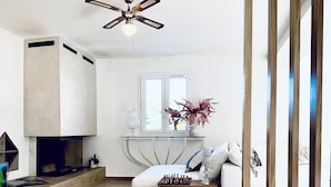 A modern living room with a fireplace, a sofa, a coffee table with a chess set, and a window with plants.