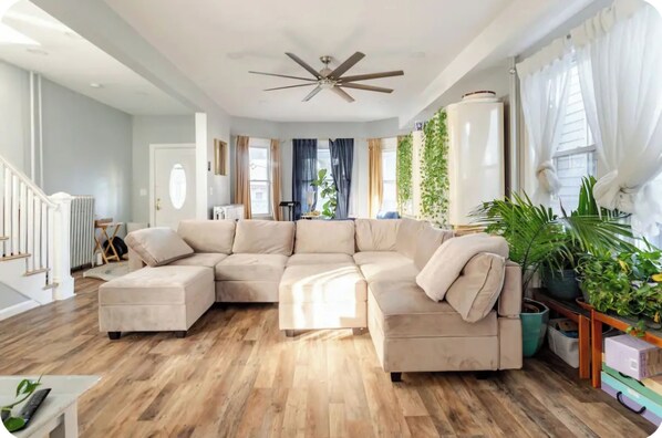 Una sala de estar moderna con un sofá modular, piso de madera, ventilador de techo y ventanas amplias con cortinas.