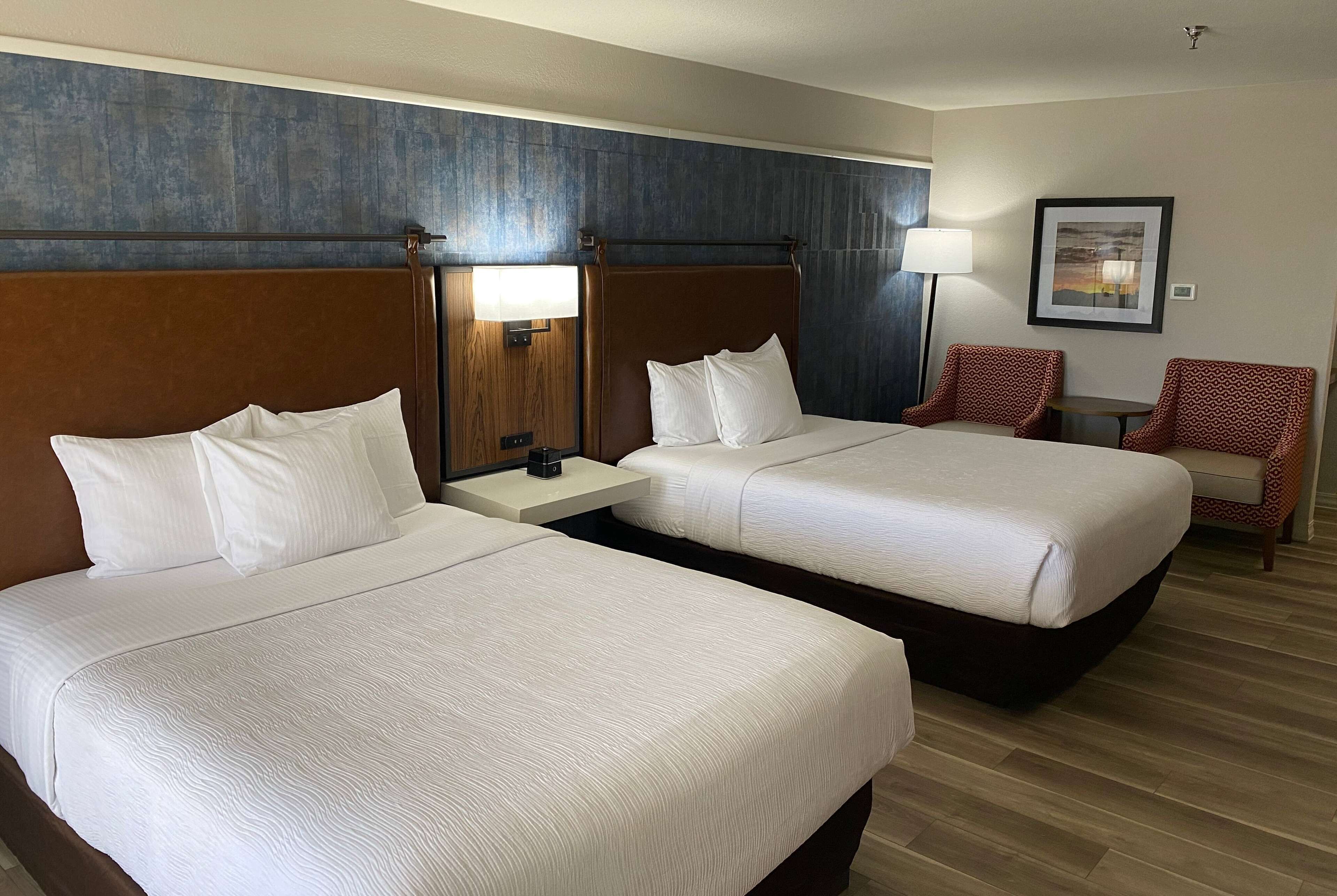 A hotel room with two beds, a nightstand, a lamp, and a chair.