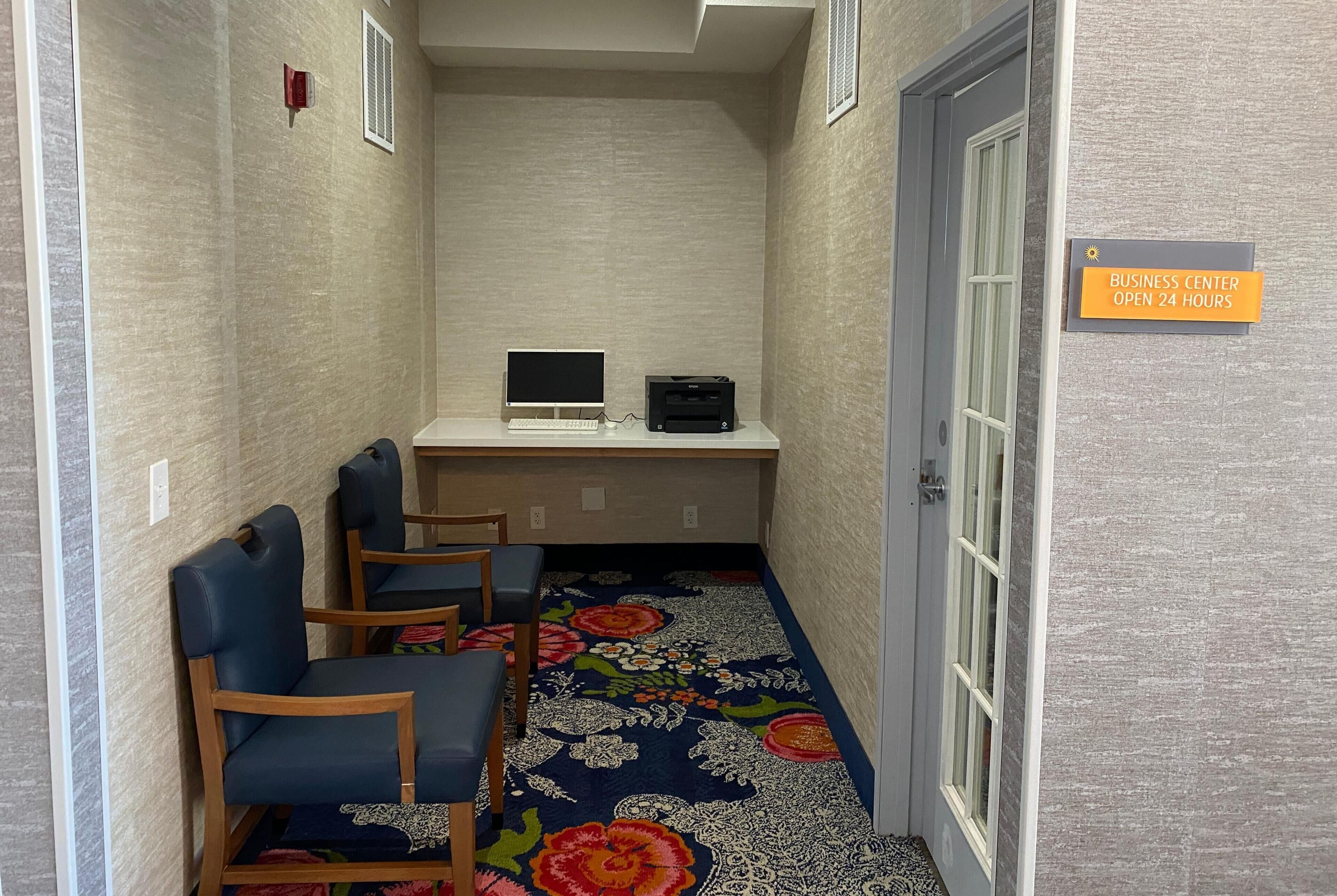Un couloir avec un bureau d’ordinateur, deux chaises et un tapis orné de motifs floraux. Une pancarte indique que le centre d’affaires est ouvert 24 heures sur 24.