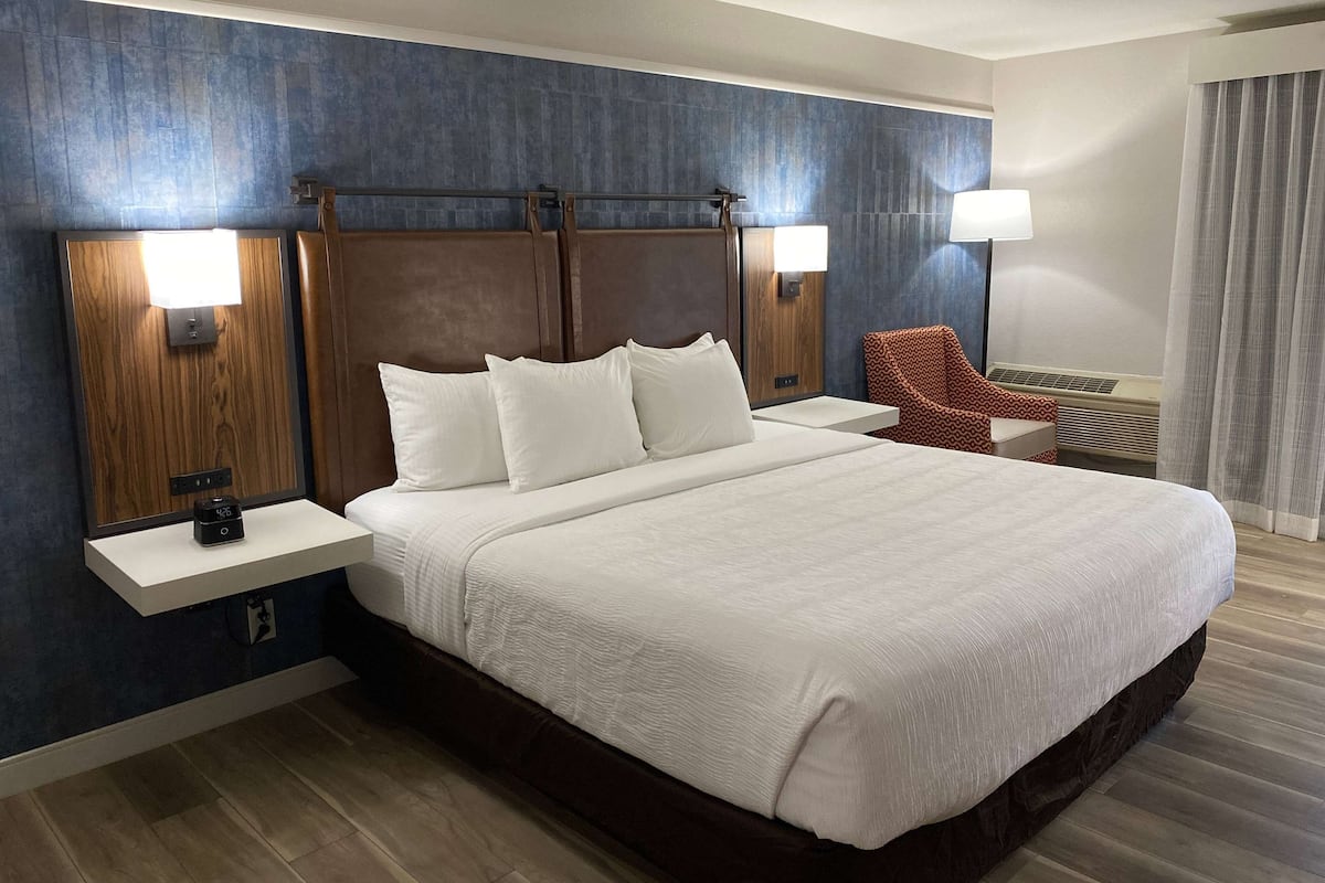 Een hotelkamer met een groot bed, twee nachtkastjes, een lamp aan de muur en een rode fauteuil met een patroon.