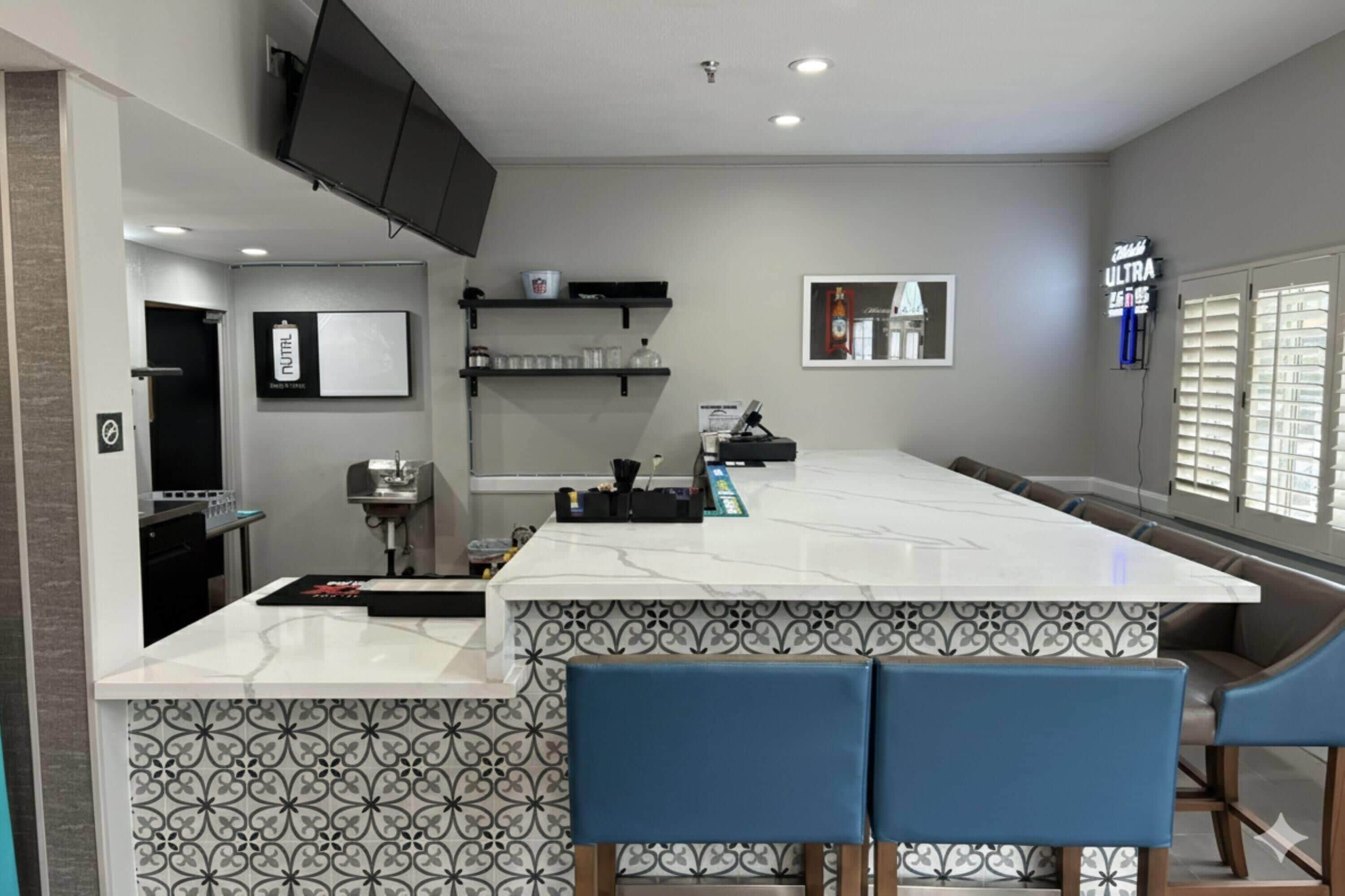 A modern kitchen with a marble countertop, blue bar stools, a flat-screen TV, and a window with blinds.