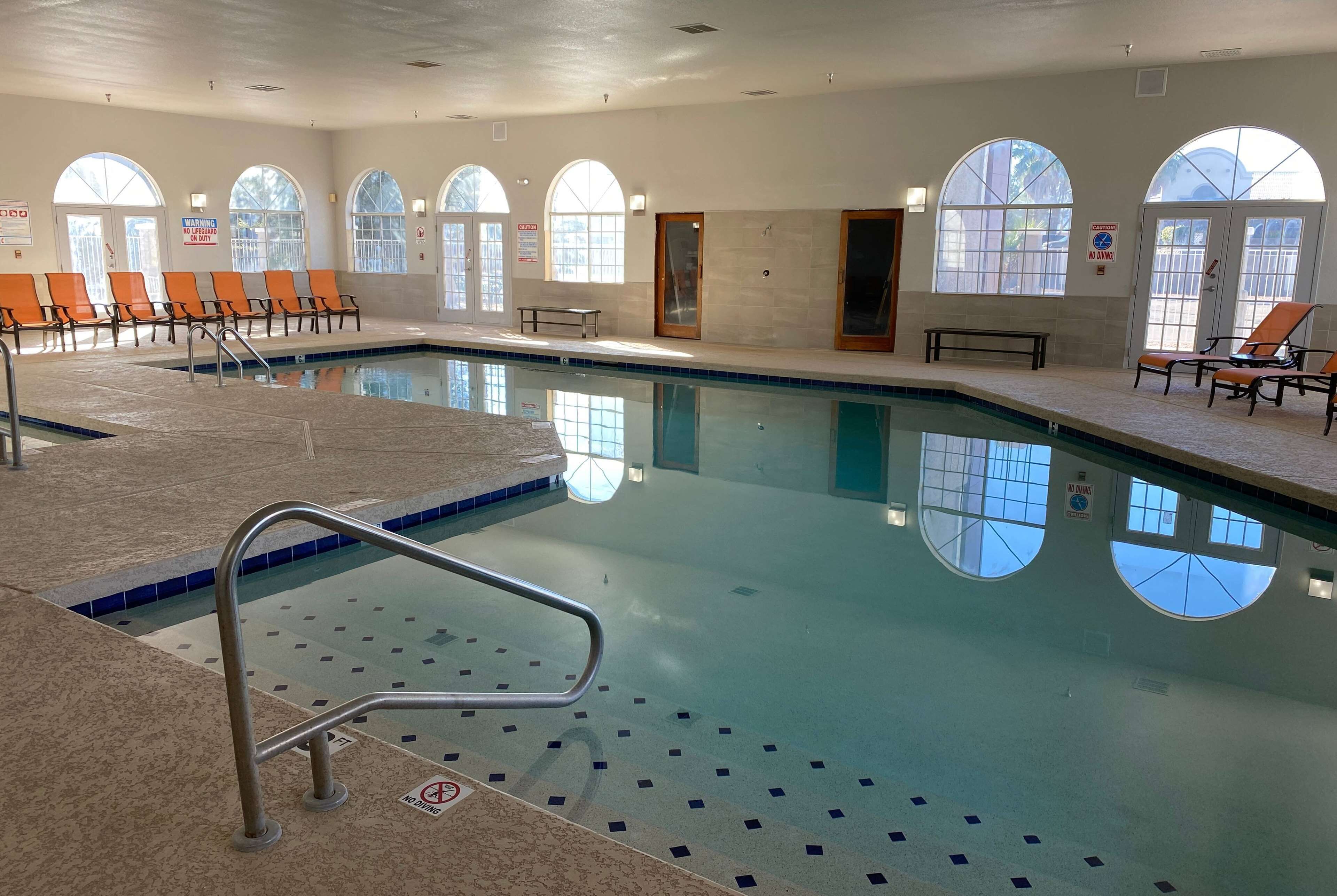 Una piscina interior con agua cristalina, iluminada por grandes ventanales arqueados, y equipada con barandales y bancos de seguridad.