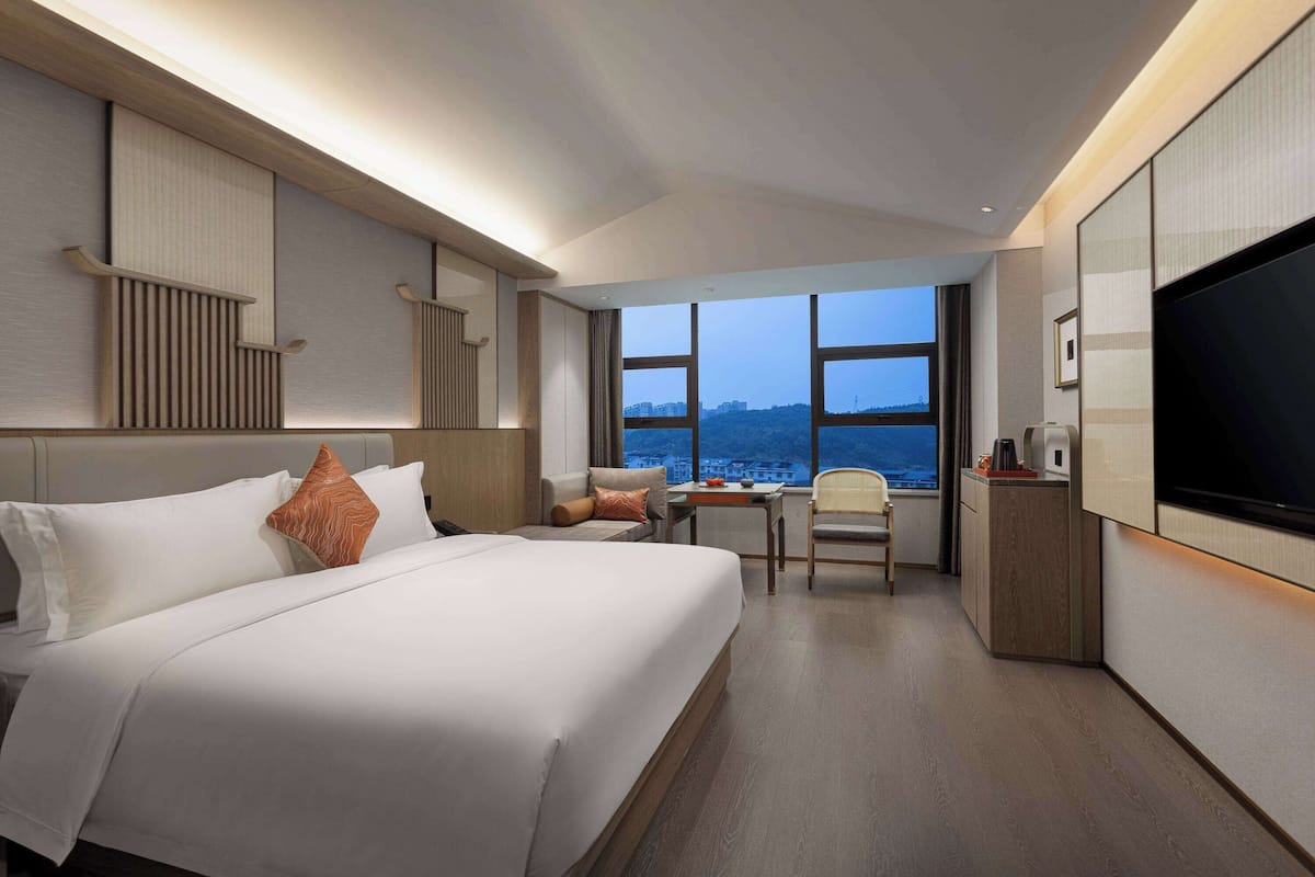 Ein Hotelzimmer mit einem großen Bett, einem Schreibtisch, einem Sessel, einem Fernseher und Blick auf die Stadt durch große Fenster.