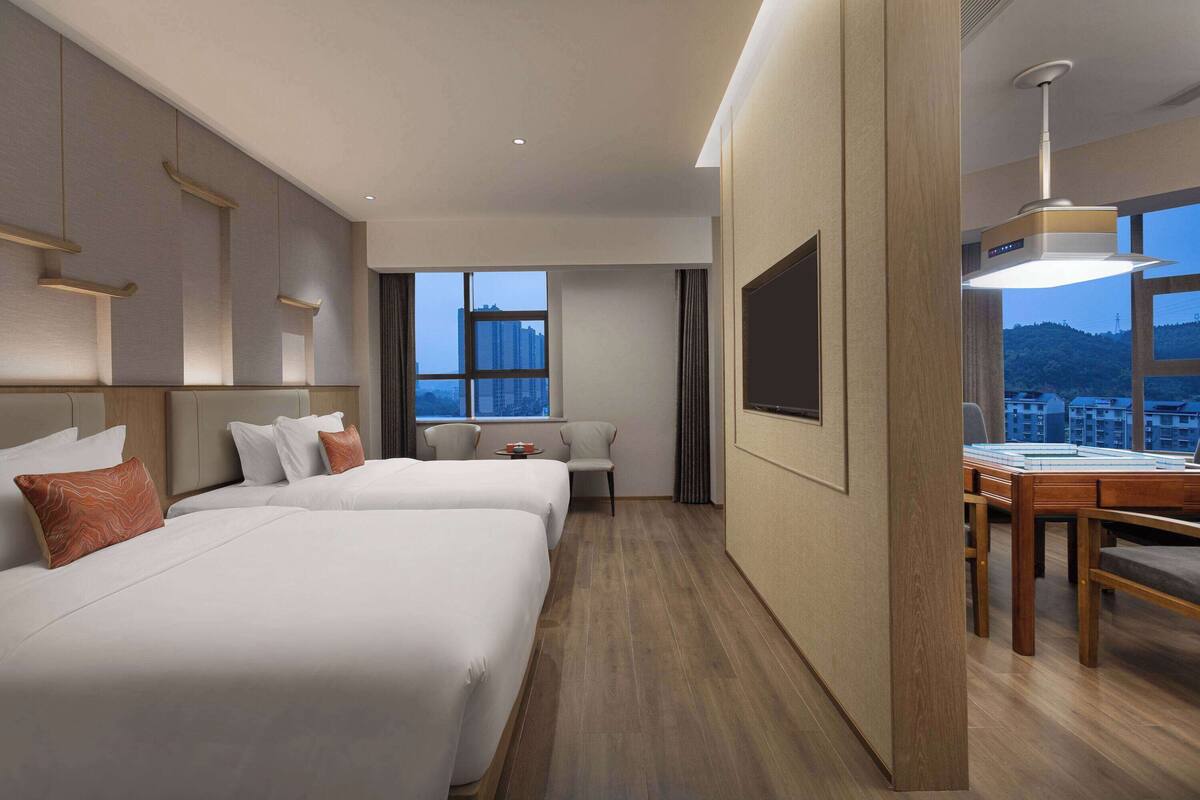 Ein Hotelzimmer mit zwei Betten, einem Schreibtisch und einem großen Fenster mit Blick auf die Stadt.
