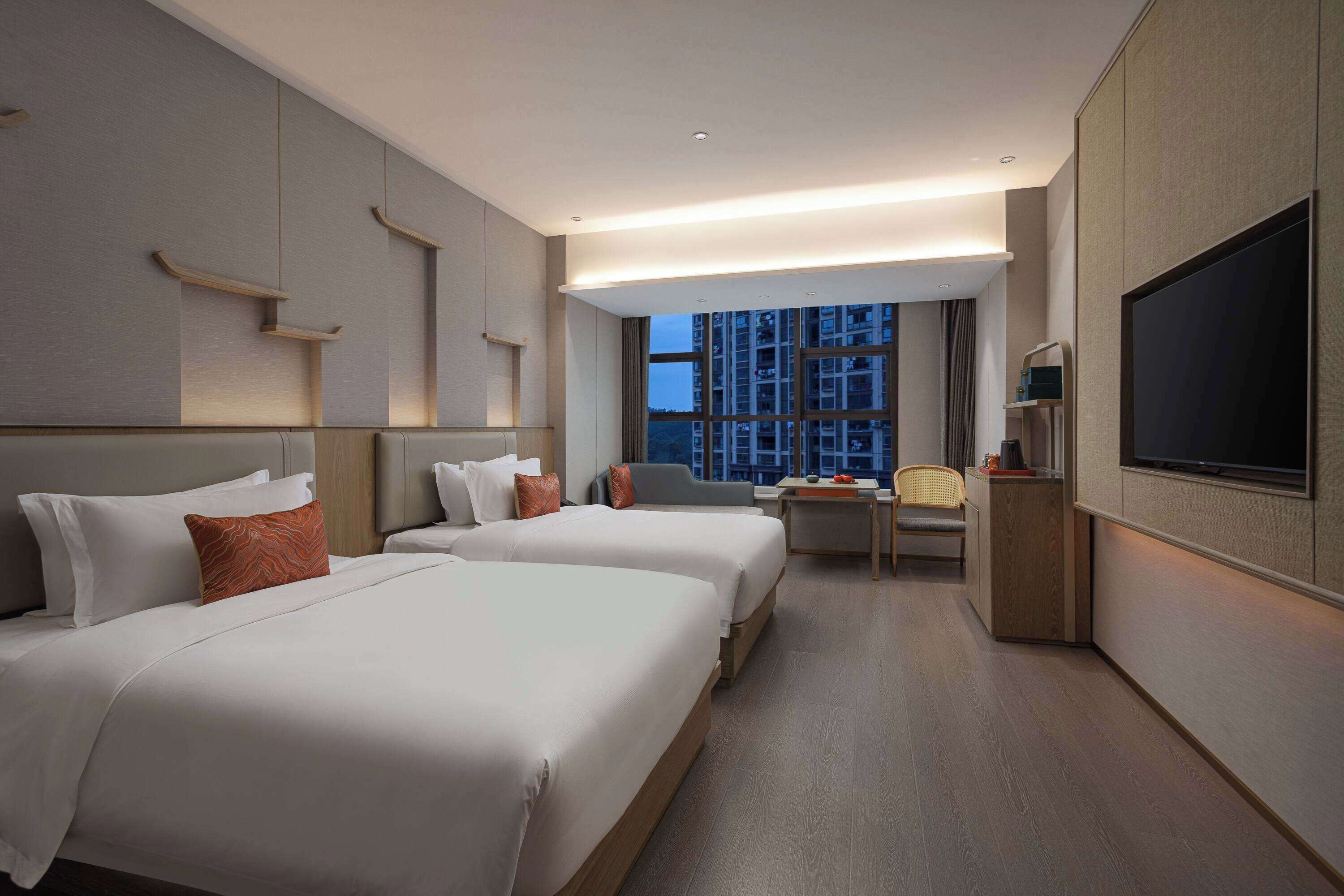 A hotel room with two beds, a desk, a chair, a TV, and a large window with a city view.