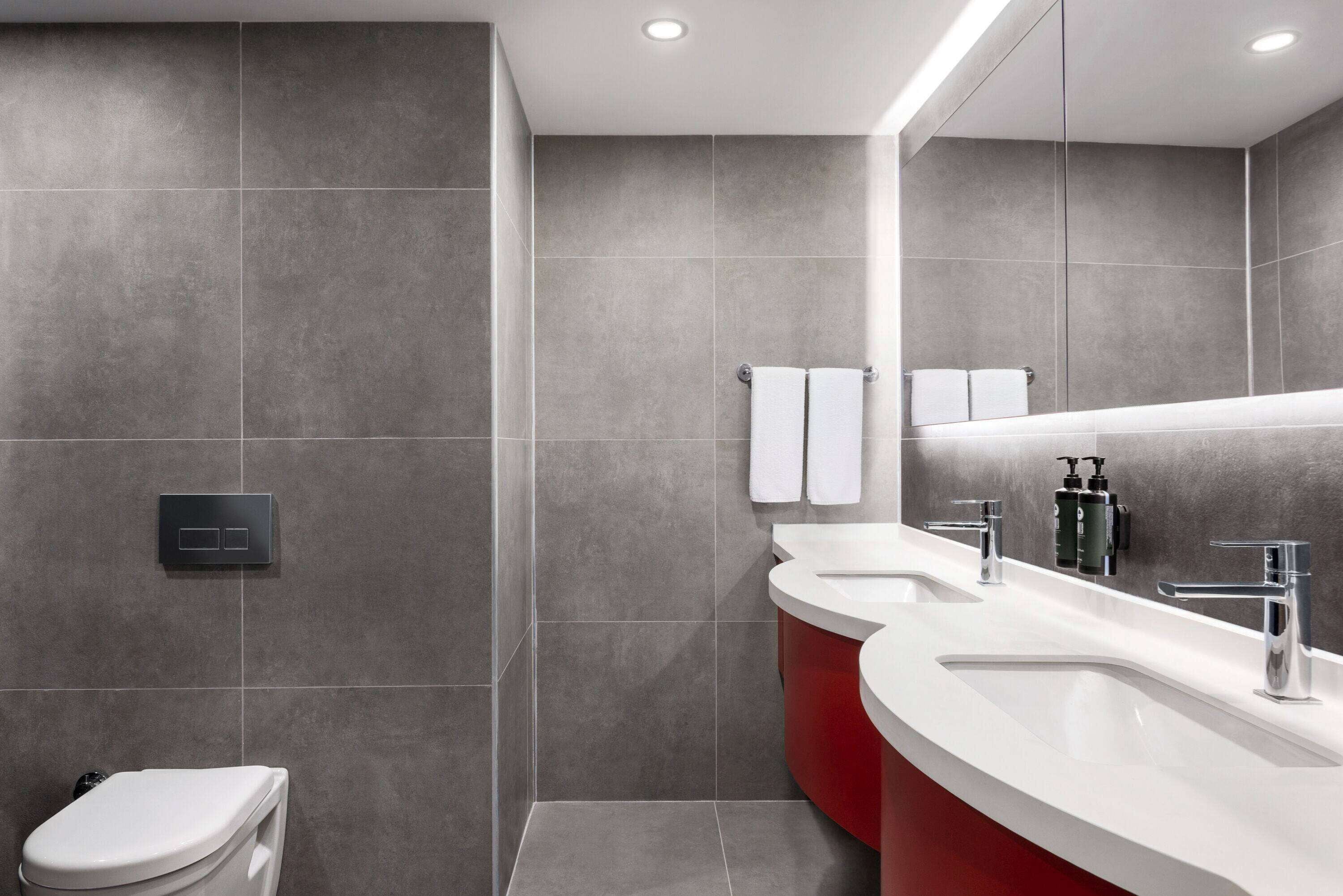 A modern bathroom with grey tiles, a white toilet, a red vanity with two sinks, and a large mirror.