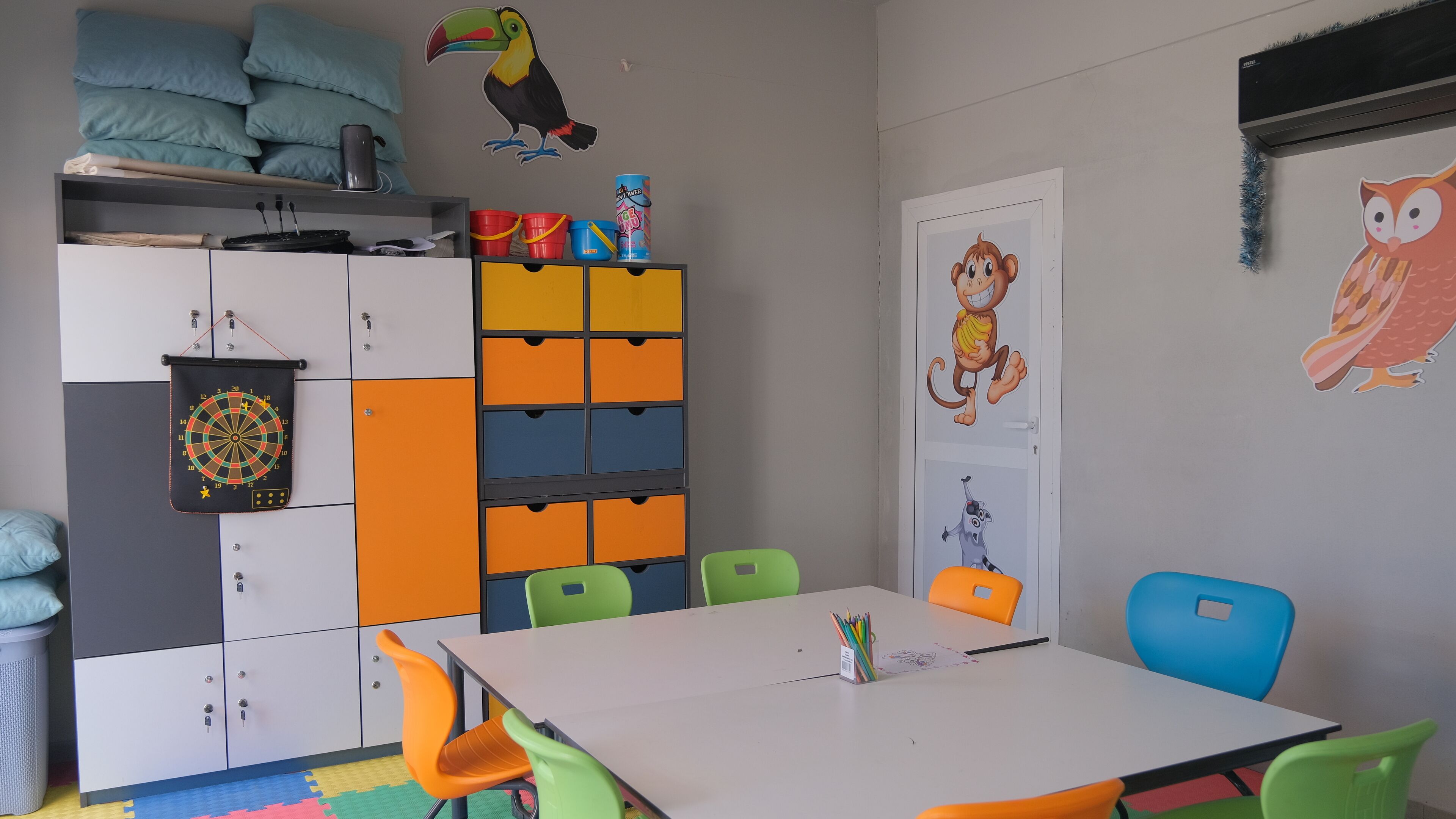 A children's room with a white table, colorful chairs, a storage unit with multiple compartments, and playful wall decorations.