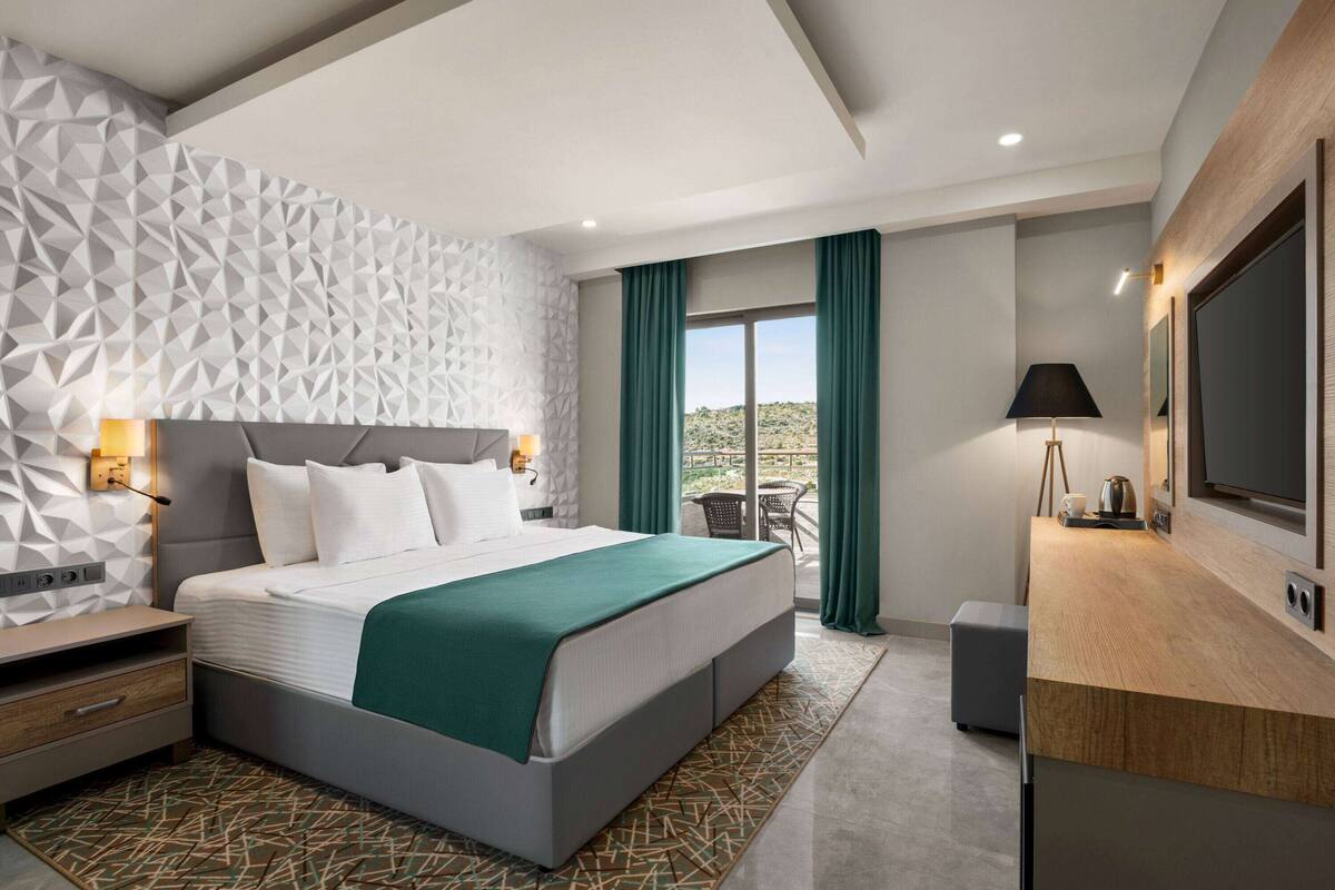 Ein modernes Hotelzimmer mit Wand in geometrischem Muster, einem großen Bett mit grüner Dekowürfe, einem Schreibtisch mit Flachbildfernseher und einem Balkon mit Blick auf die Wüste.