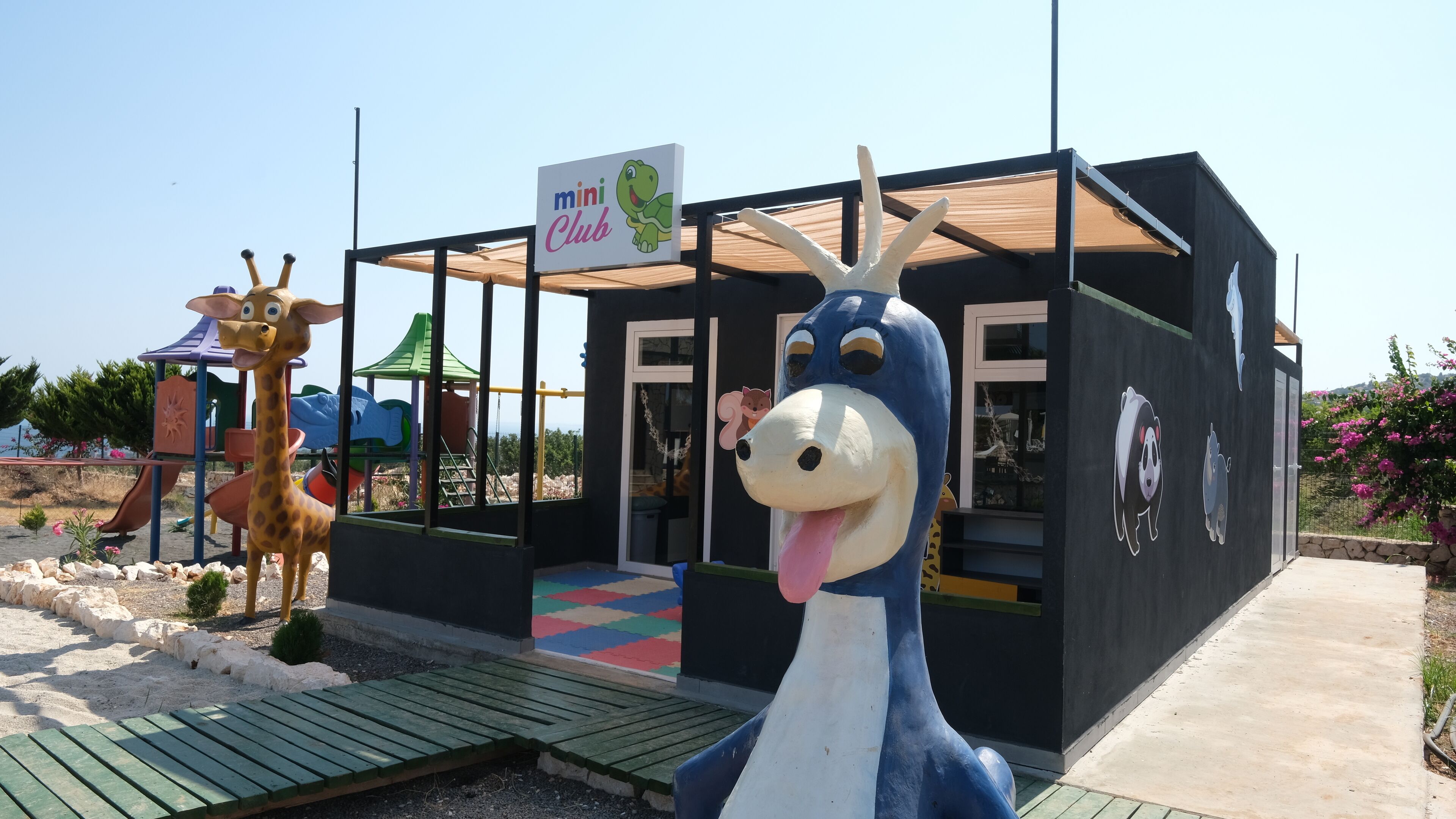 Un espace de jeux pour enfants avec une statue de girafe et une statue d’éléphant bleu, un panneau indiquant « mini-club », et une maison de jeux avec un toit coloré.