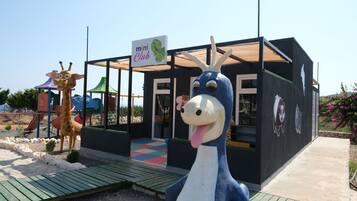 Un espace de jeux pour enfants avec une statue de girafe et une statue d’éléphant bleu, un panneau indiquant « mini-club », et une maison de jeux avec un toit coloré.