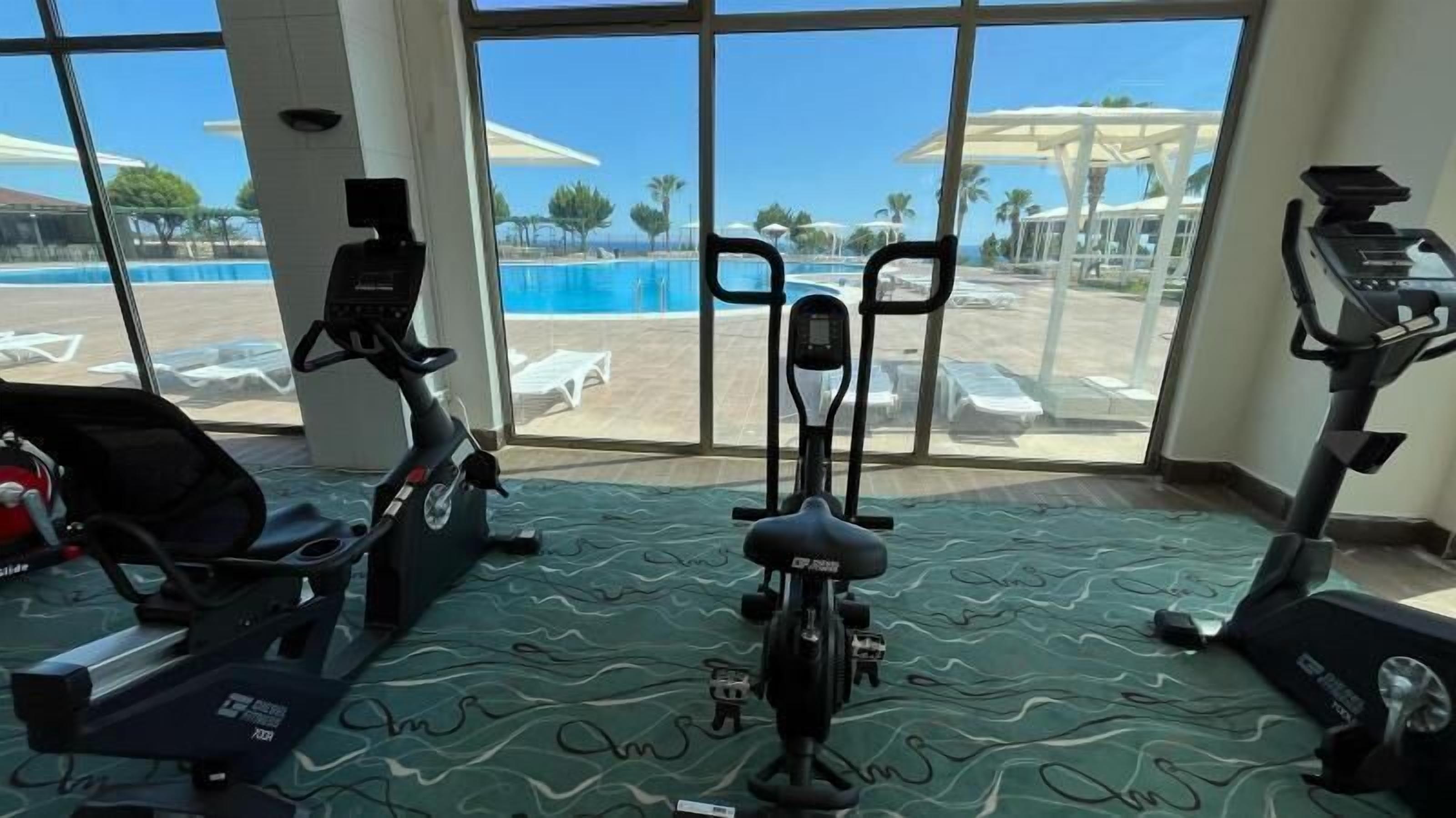 A gym with exercise equipment and a view of a pool and beach through large windows.