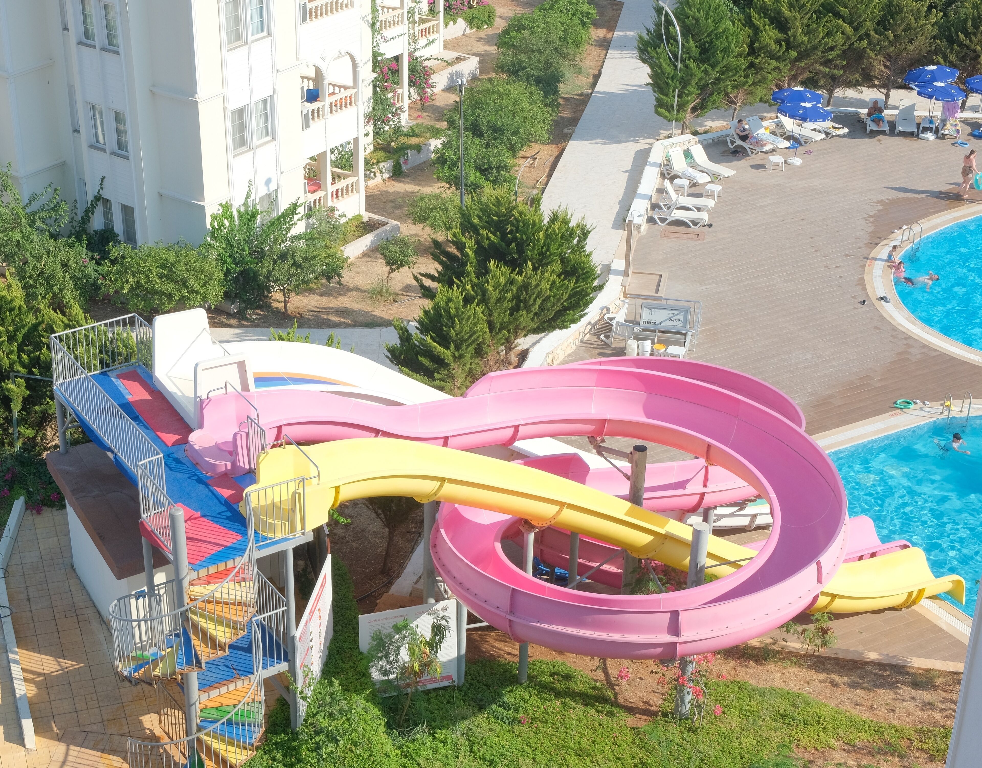 A water park with a large multi-colored water slide, a pool area with lounge chairs, and a building with balconies.