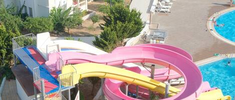A water park with a large multi-colored water slide, a pool area with lounge chairs, and a building with balconies.