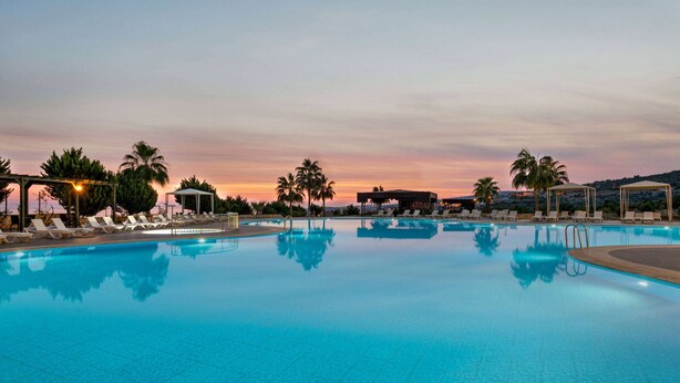 A large outdoor swimming pool with multiple lounge chairs and palm trees, surrounded by a covered area with a sunset view.