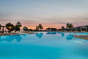 Una amplia piscina exterior con numerosas tumbonas y palmeras, rodeada por una zona cubierta con vistas al atardecer.