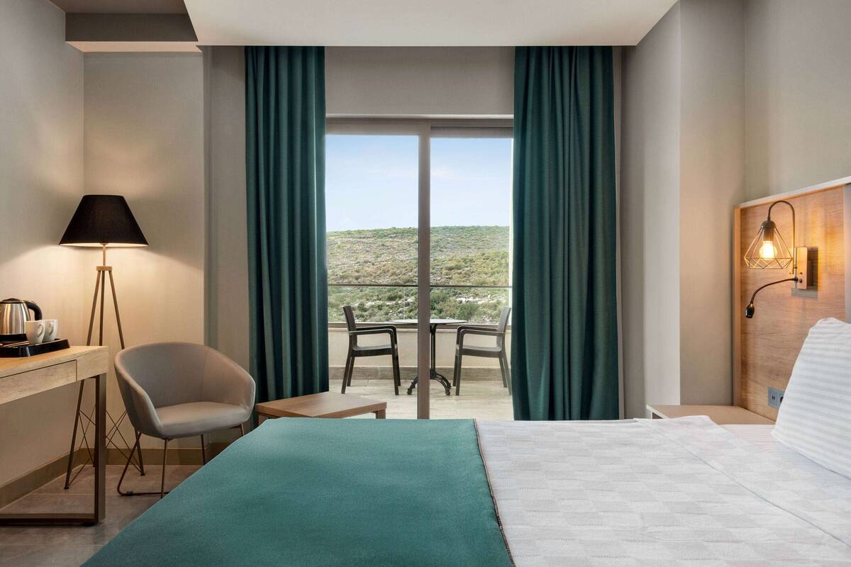 Ein Hotelzimmer mit einem Bett, einem Schreibtisch mit Lampe, einem Sessel, einem kleinen Tisch und Blick auf eine Wüstenlandschaft durch große Fenster.