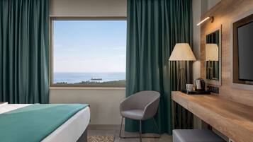 A hotel room with a bed, a chair, a desk with a lamp, a television, and a view of the sea.