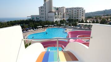 A view of a swimming pool with a water slide, surrounded by lounge chairs and umbrellas, with a building in the background.