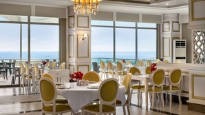 A dining area with tables set for service, chairs, a chandelier, and a view of the sea.