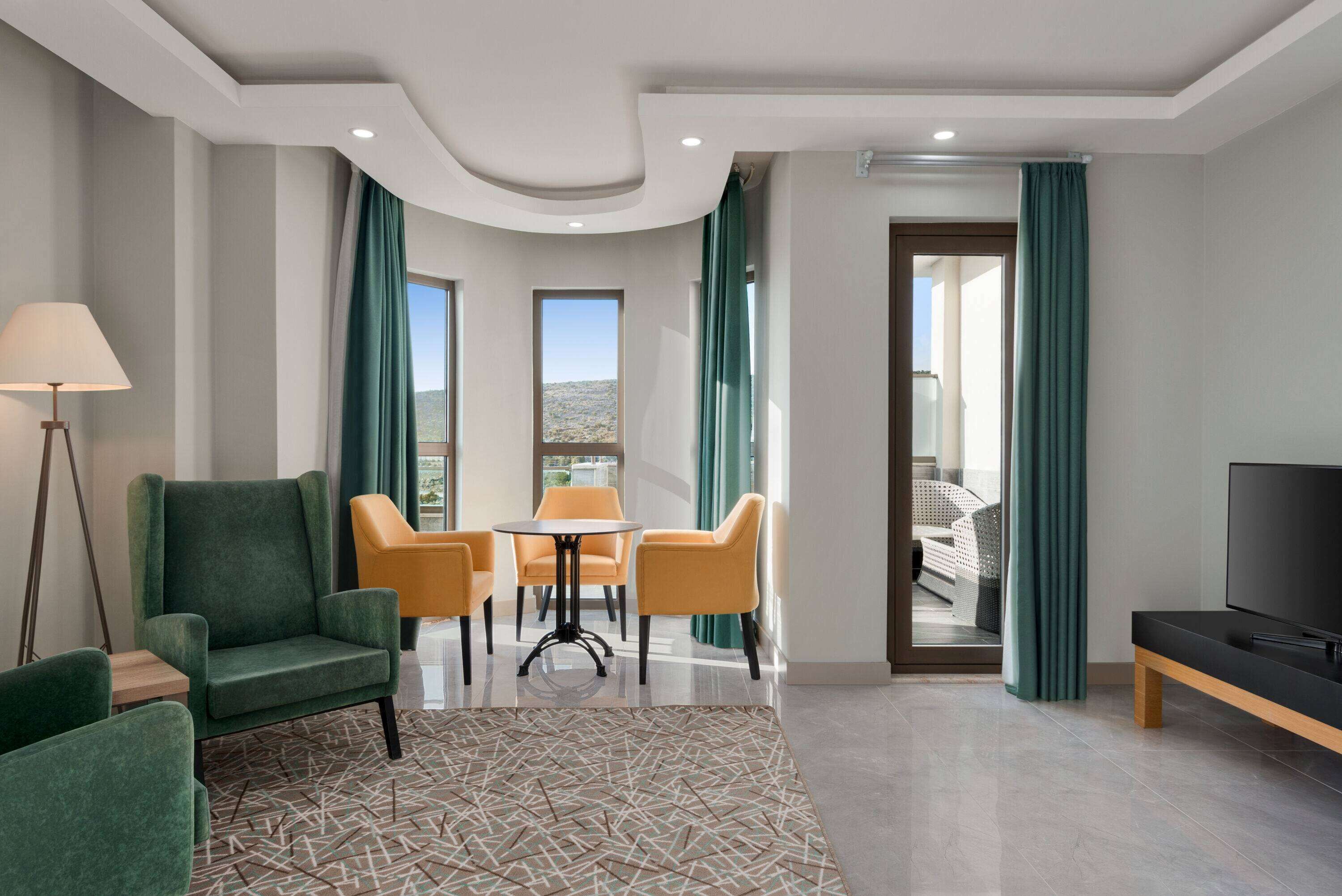 A hotel room with a green armchair, a small table, a sofa, a TV, and a balcony with a view of the mountains.