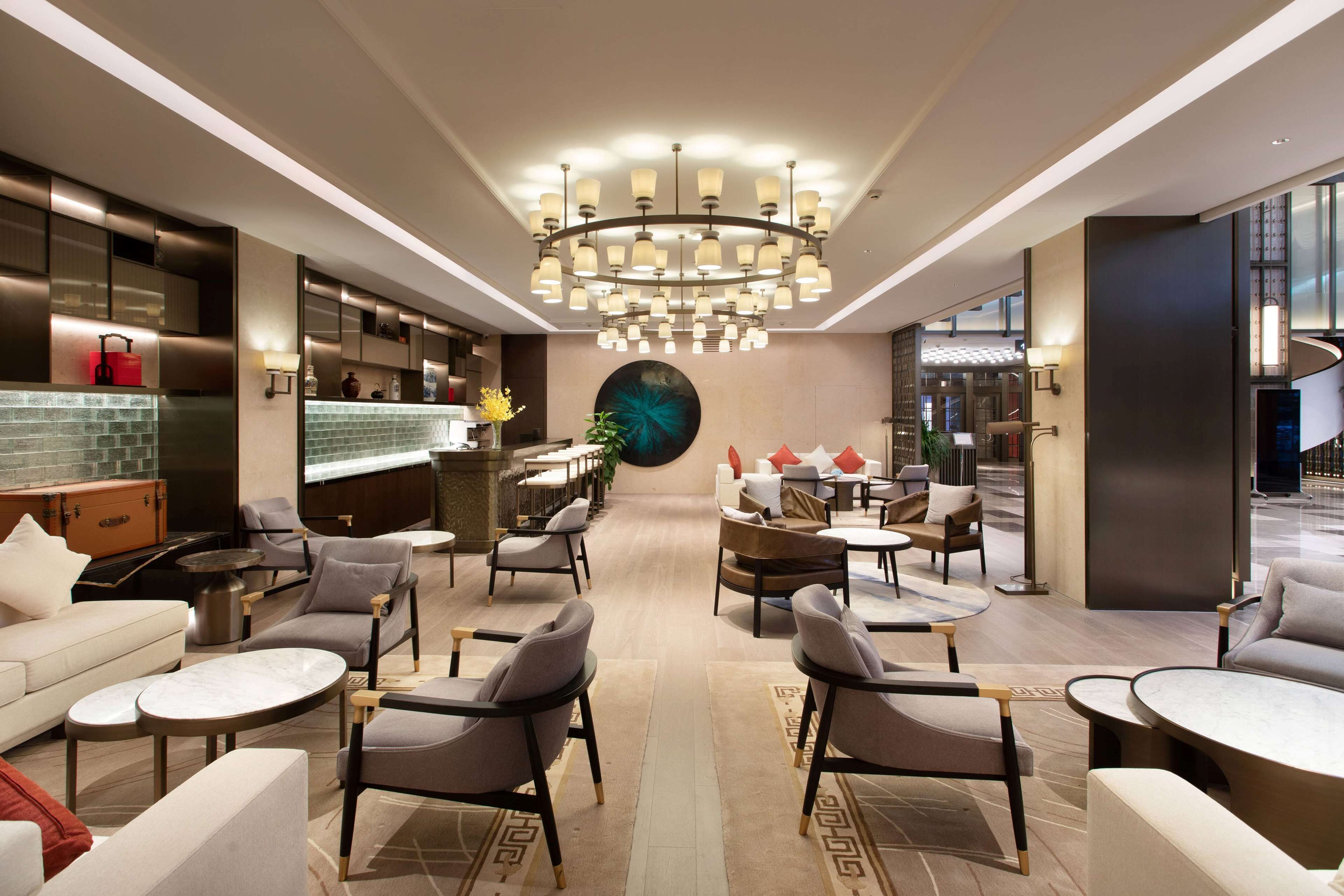 Un hall d'hôtel moderne avec un lustre central, des espaces de détente et un bar.