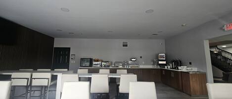 Un comedor moderno con mesas y sillas blancas, una encimera de cocina con electrodomésticos y una escalera que sube a otro nivel.
