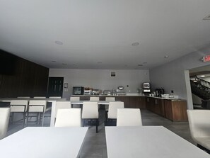 Un comedor moderno con mesas y sillas blancas, una encimera de cocina con electrodomésticos y una escalera que sube a otro nivel.