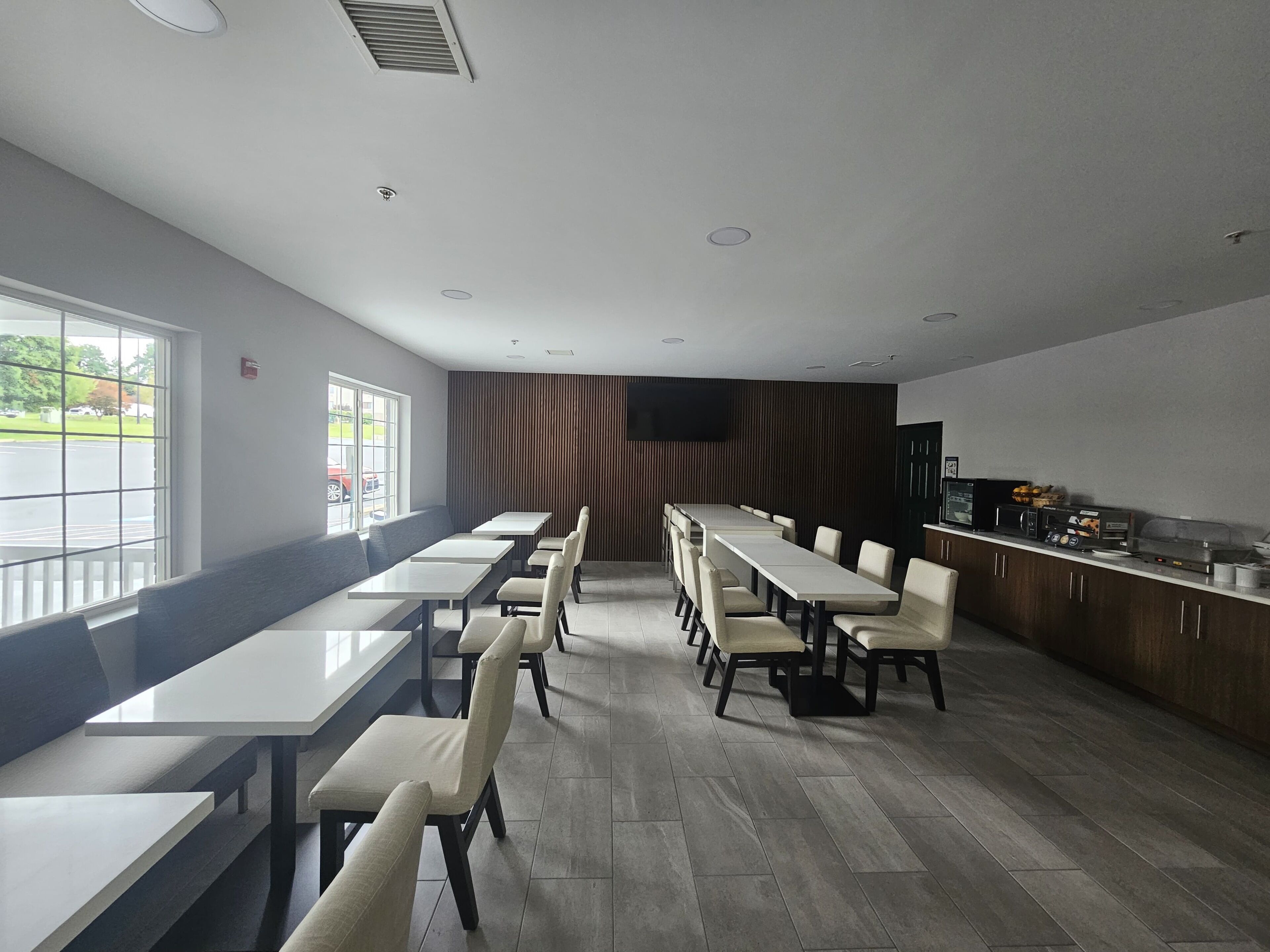 A modern dining area with long tables and chairs, a kitchen counter with appliances, and a large window providing a view of the outdoors.