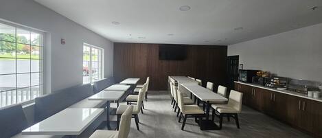 A modern dining area with long tables and chairs, a kitchen counter with appliances, and a large window providing a view of the outdoors.