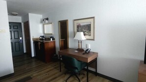 Una habitación peque ña y de planta libre con un escritorio de madera, una silla, una lámpara y un cuadro en la pared.