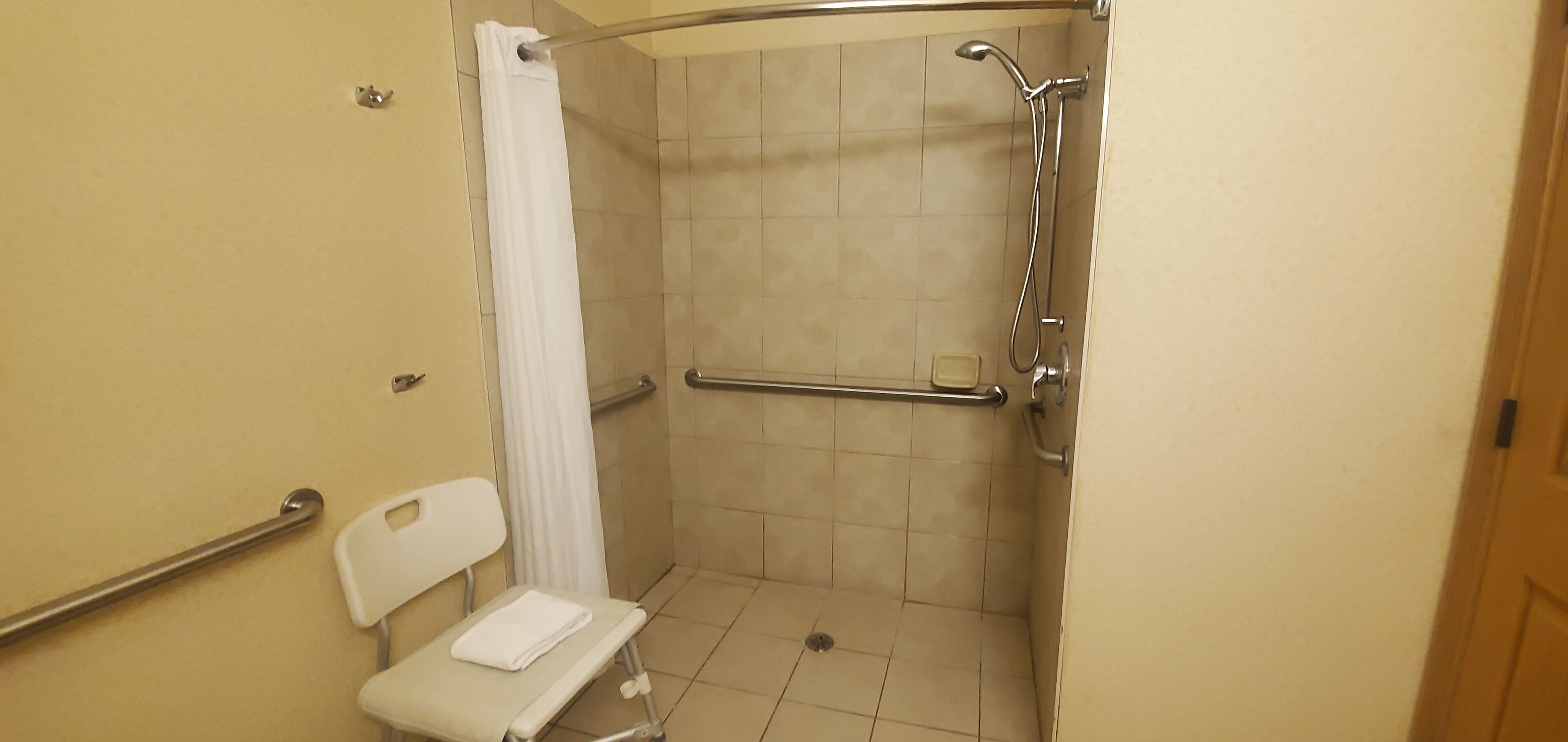 A wheelchair-accessible shower stall with a handrail, a white chair with a towel, and a handheld showerhead.