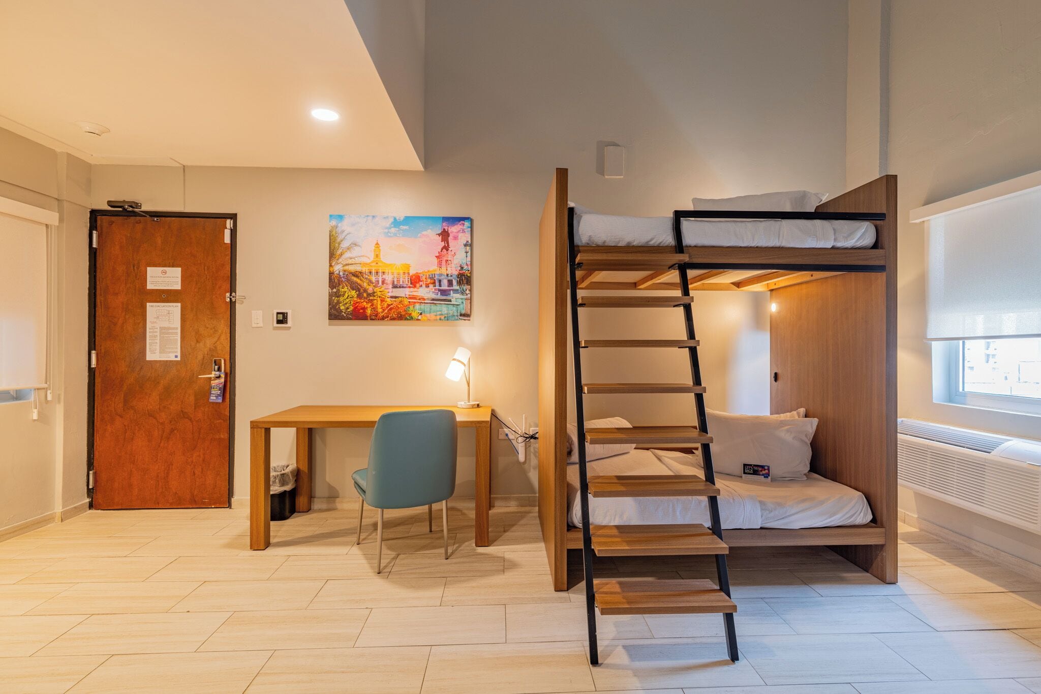 A room with a bunk bed, a desk with a chair, a painting on the wall, and a door with a notice.