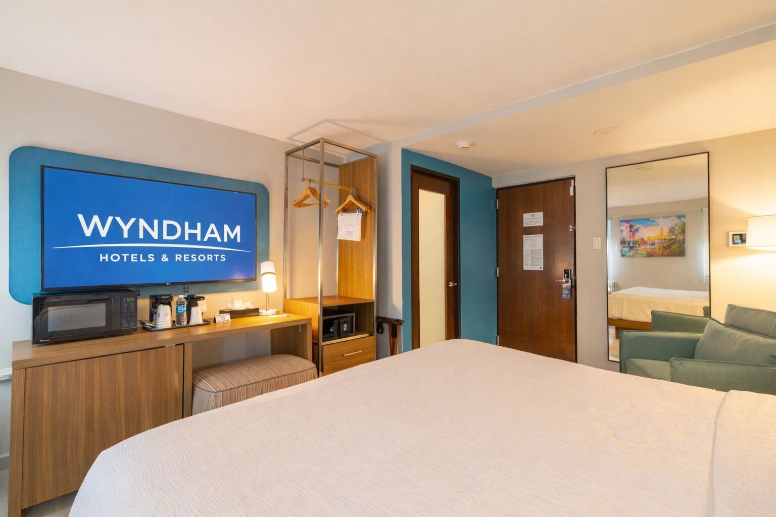 Habitación de hotel con cama, televisor mostrando 'WYNDHAM HOTELS & RESORTS', escritorio con microondas, cafetera y perchero.