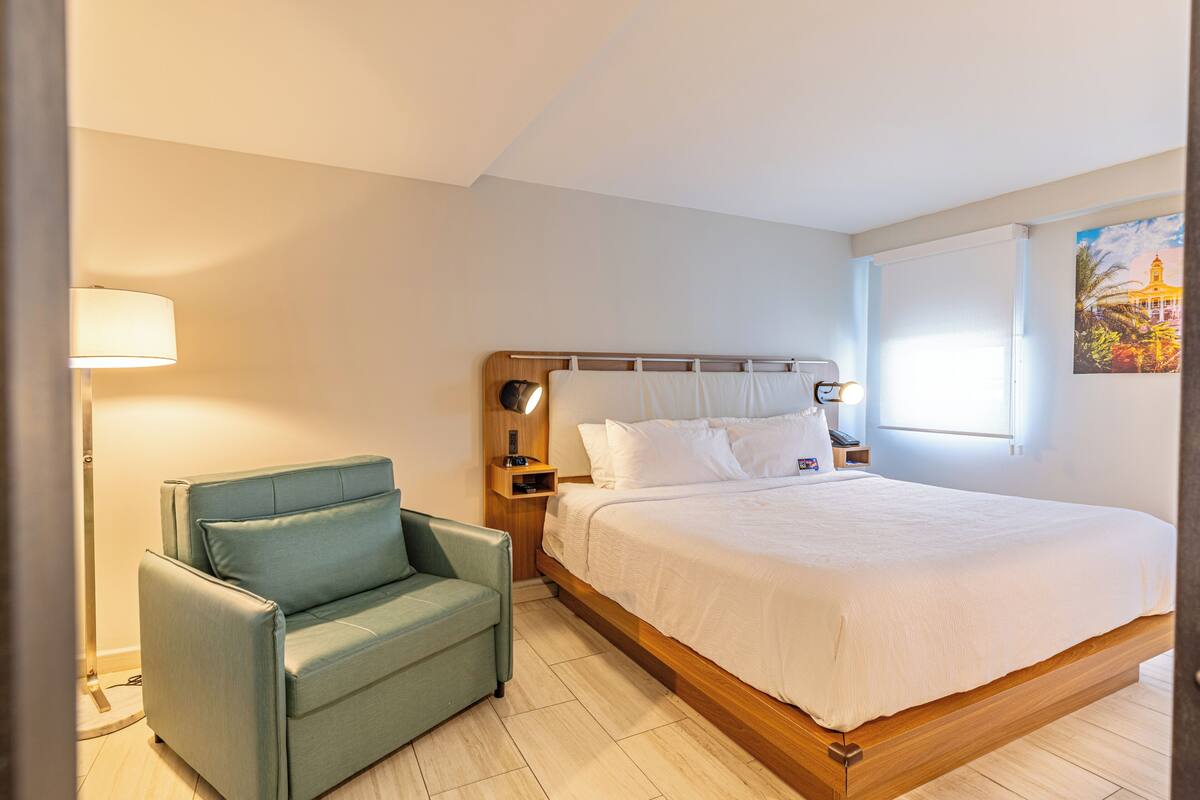 A bedroom with a bed, a nightstand, a lamp, a chair, and a painting on the wall.