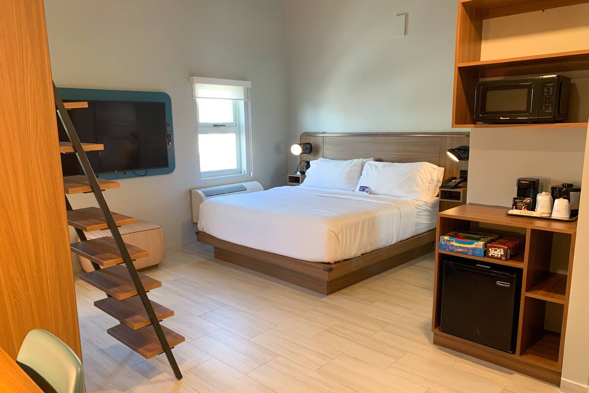 A bedroom with a bed, a small refrigerator, a microwave, a desk with a computer, and a window.