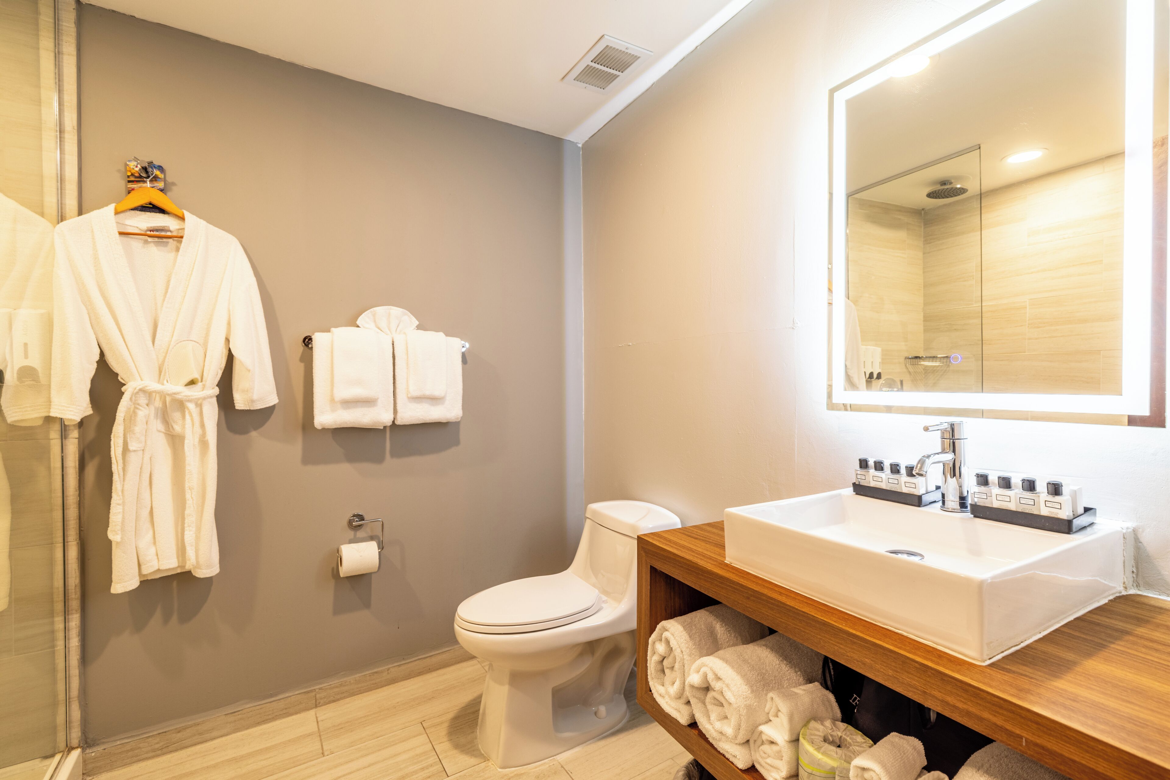 A modern bathroom with a toilet, sink, and mirror. There are towels and robes hanging on the wall.