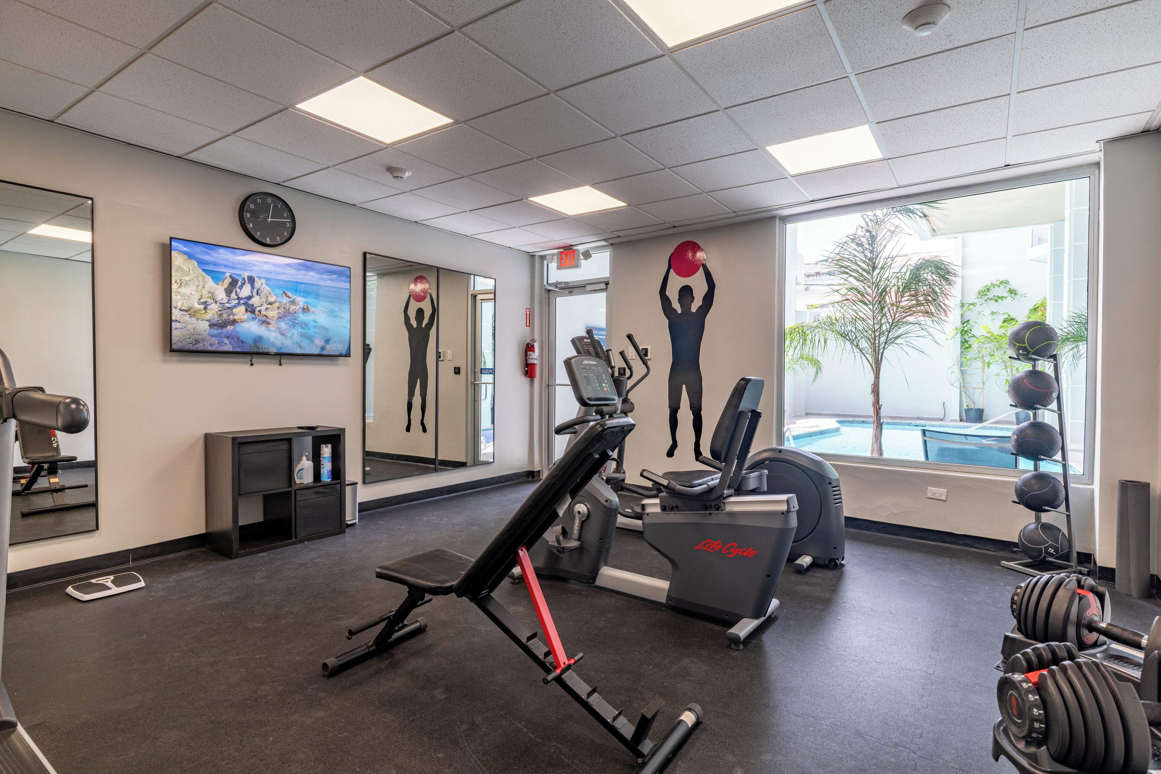 A gym with exercise equipment, a large mirror, a clock, and a painting of a rocky coastline.
