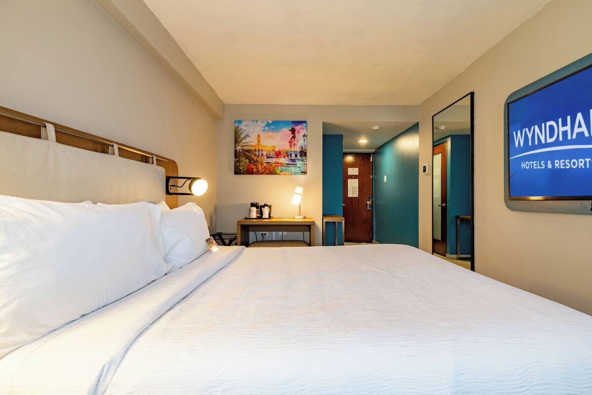 A hotel room with a bed, a desk with a lamp, a painting on the wall, and a sign for Wyndham Hotels & Resorts.