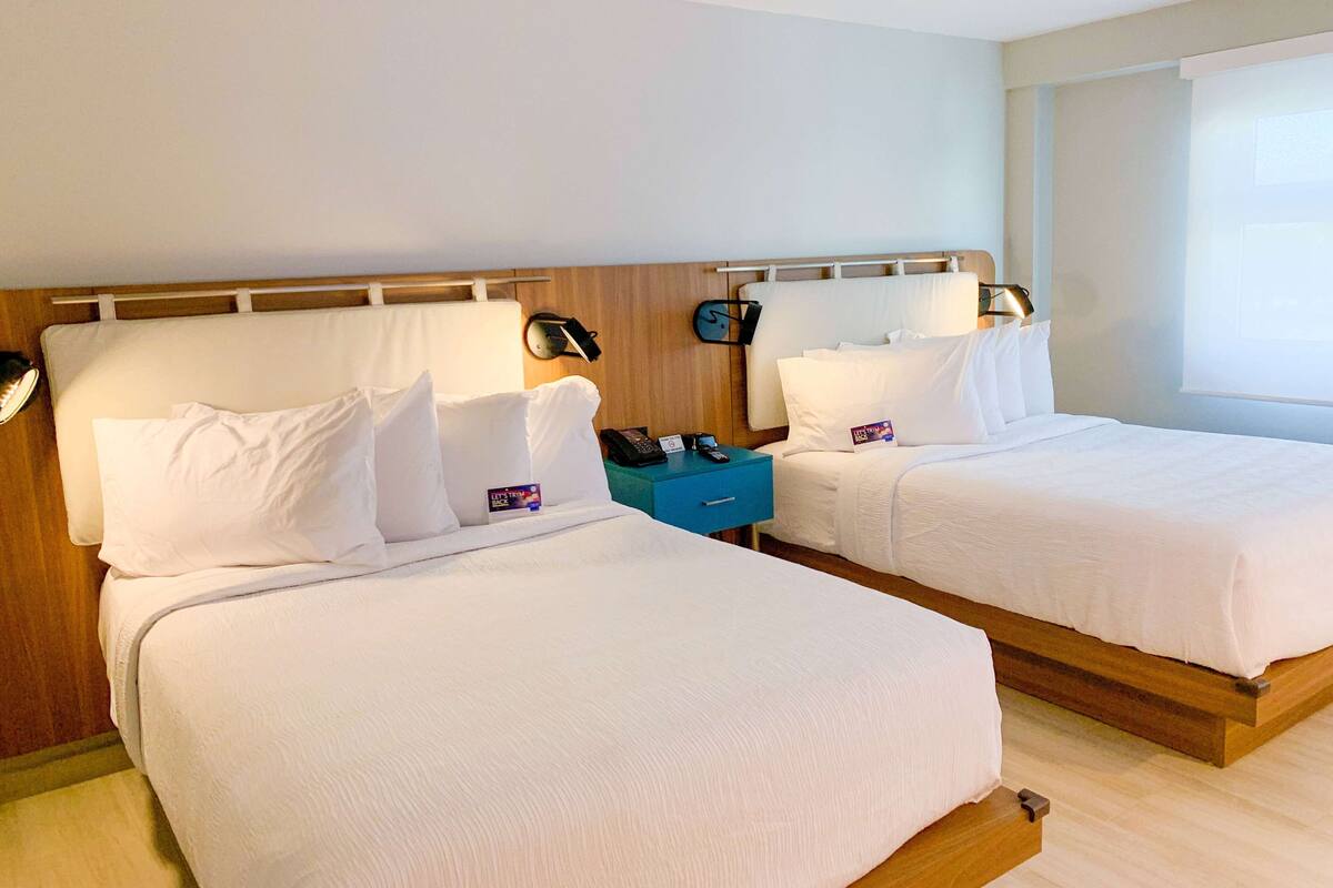 A hotel room with two beds, wooden headboards, white bedding, a blue nightstand, and a bedside table with a lamp.