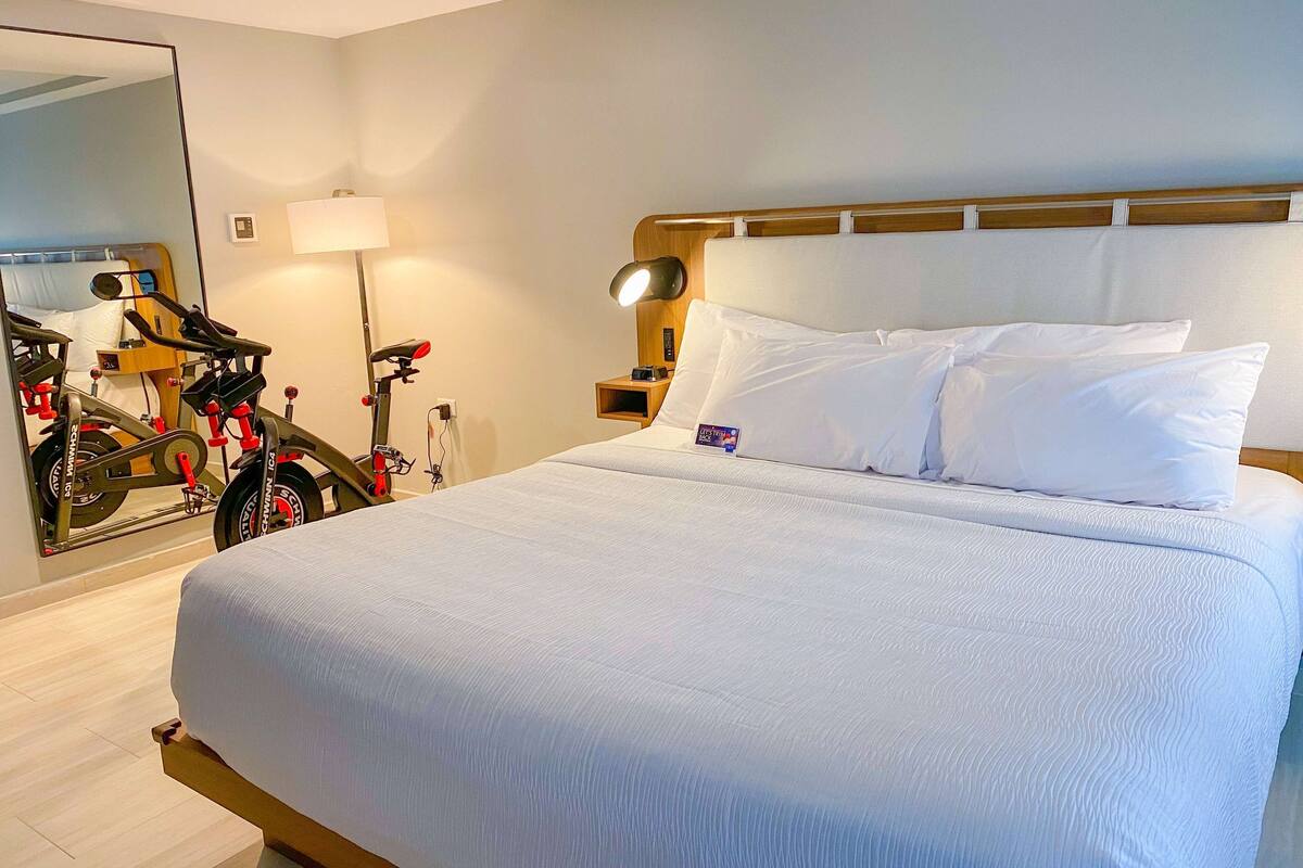 A hotel room with a large bed, a bicycle in the corner, a nightstand with a lamp, and a mirror reflecting the room.